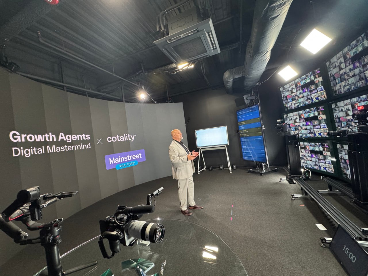 Wide view of Matt presenting on a broadcast stage with professional cameras and a wall of remote attendees